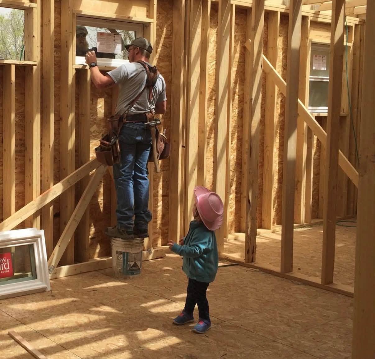 Builder installing a window on a framed home while a young child in a pink cowboy hat watches